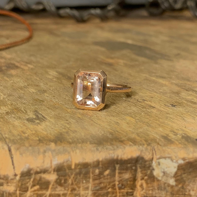 Ein rotgoldener Ring mit einem 8-eckigen, facettierten Morganit liegt auf einer Holzplatte